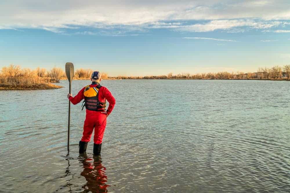 A man on a lake with paddle wearing a drysuit, lifejacket, and boots.