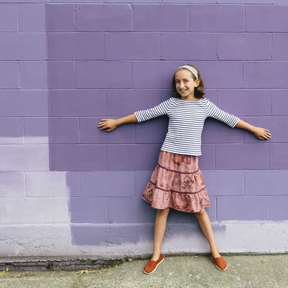 Girl in a tiered skirt stretching her arms against the purple brick wall.