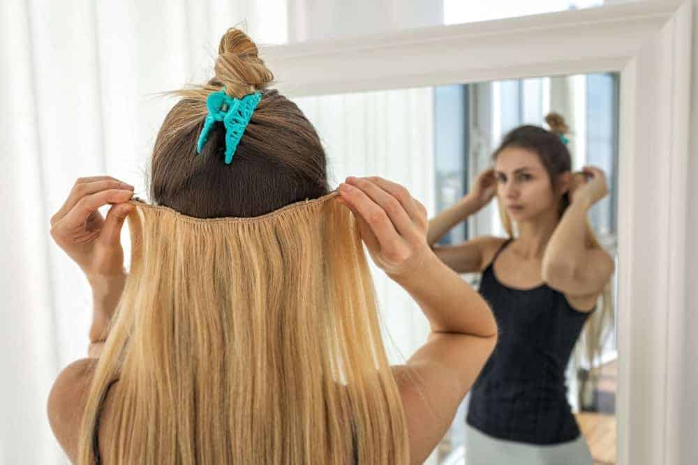 A woman putting on a blonde hair extension in front of a mirror.