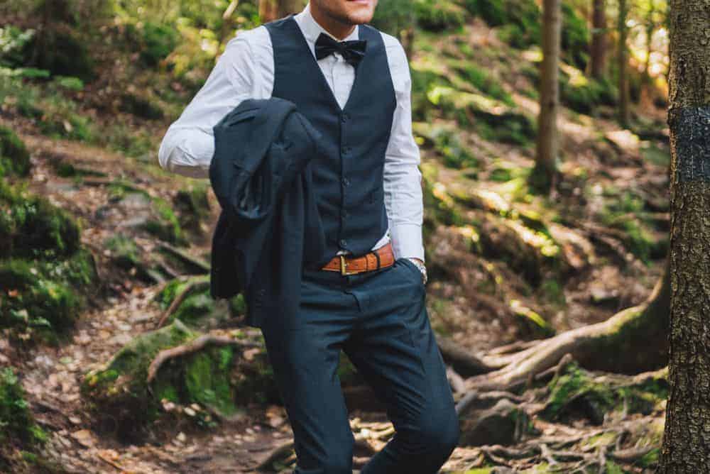Groom in the middle of a forest wearing three piece black wedding suit with a bowtie.