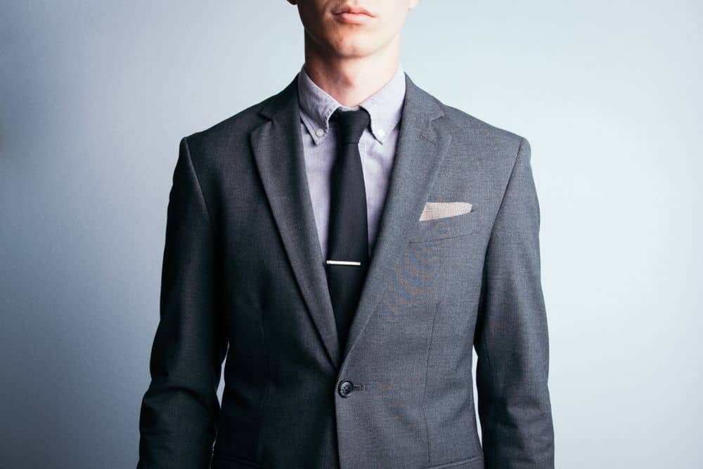 A young man sports a two piece, grey suit with a tie and pocket square.