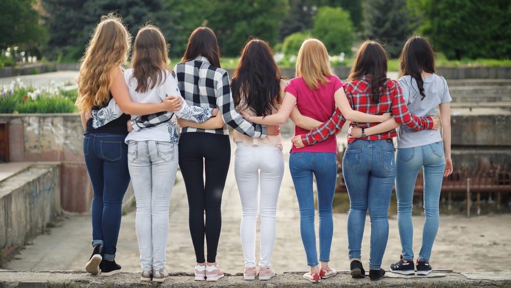 Back profile of a group of female friends in pants and shirts hugging each other.