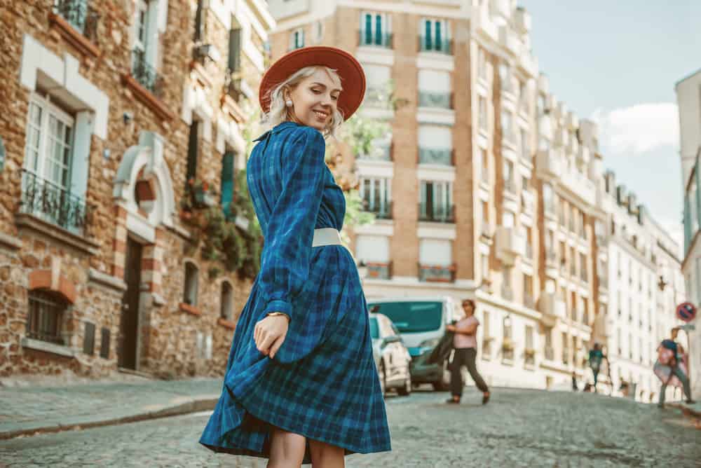Woman in a blue checkered dress with belt and hat walking along the street of Paris.