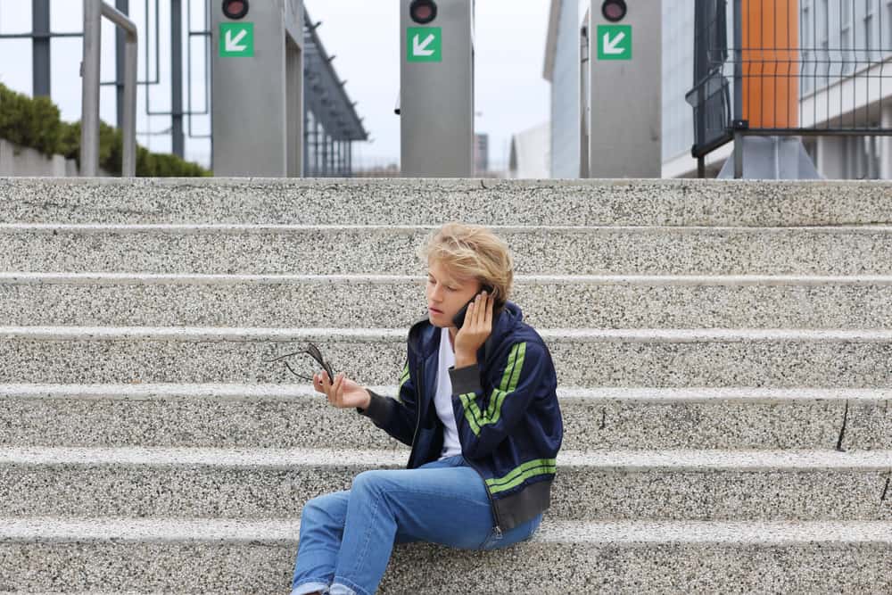 Teenage boy sitting on a city street with his phone and glasses.