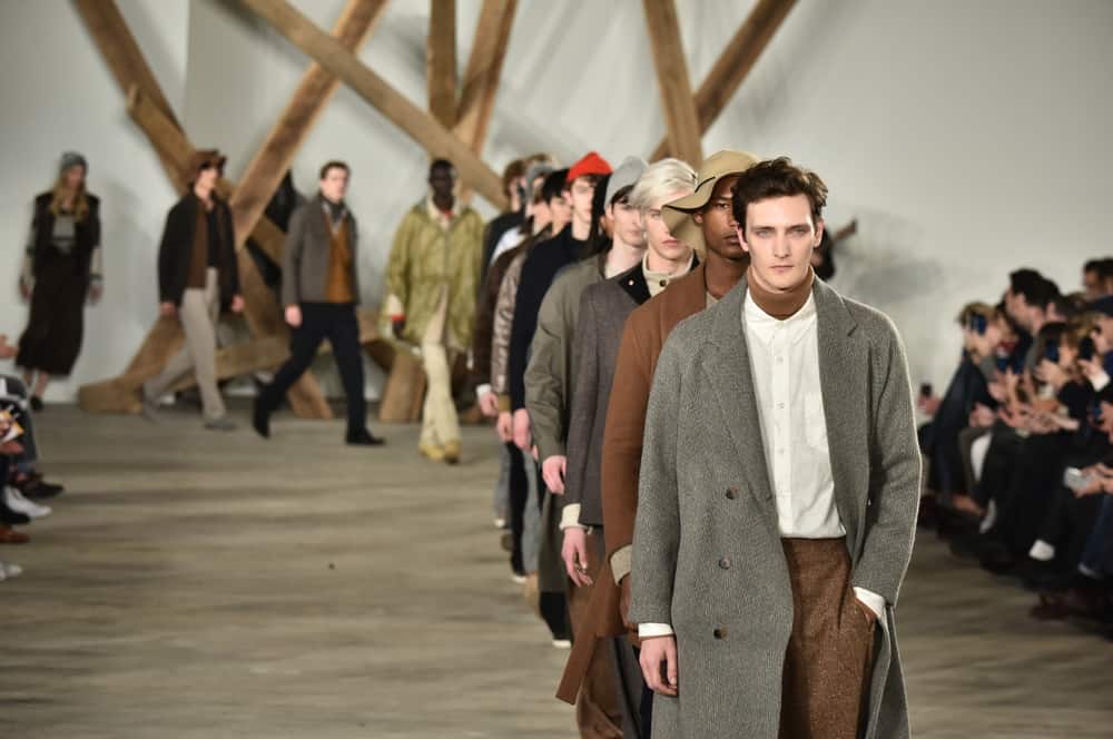 Male models in trench coats walk the runway.