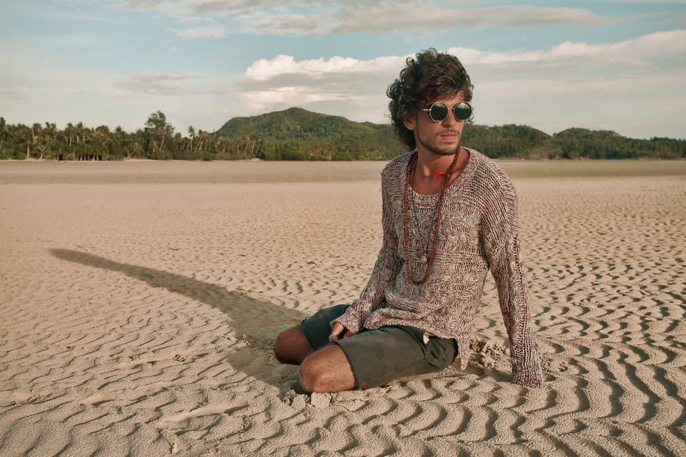 A hippie in sunglasses sitting on the beach sand.