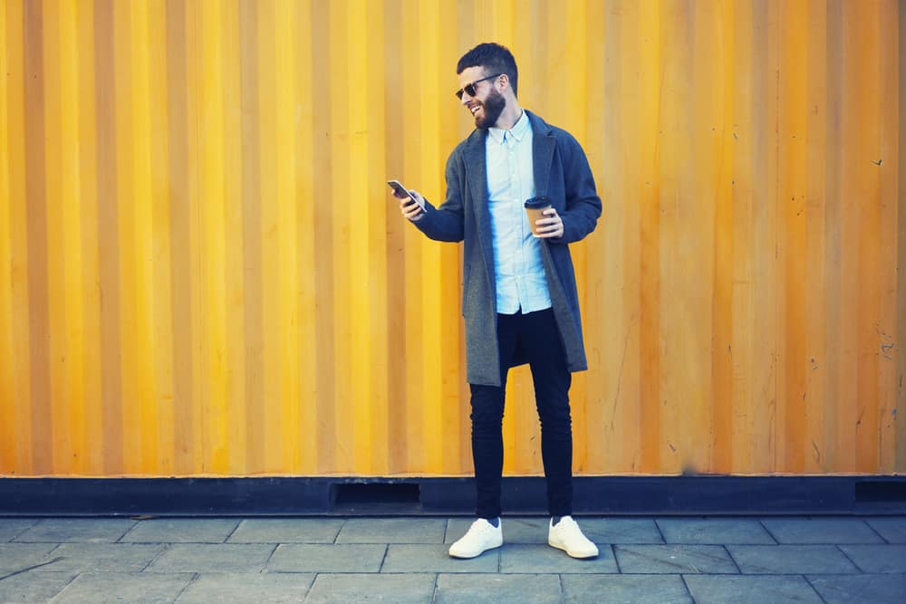 Bearded hipster holding a cup of coffee while using his phone on the other hand.