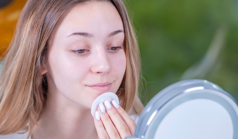 Woman removing makeup using a cotton pad in front of a mirror.