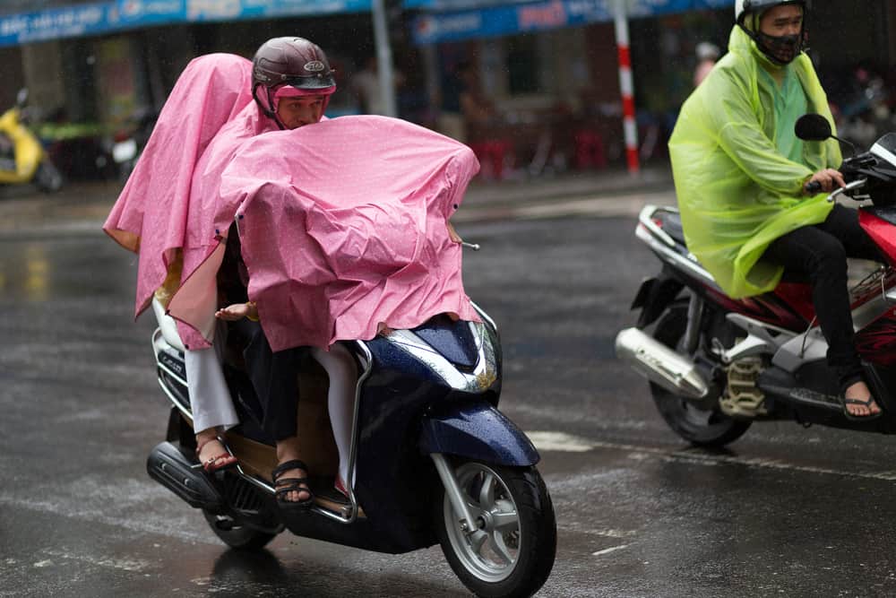 A family of three riding on a motorcycle through the rain covered with a pink tarp.