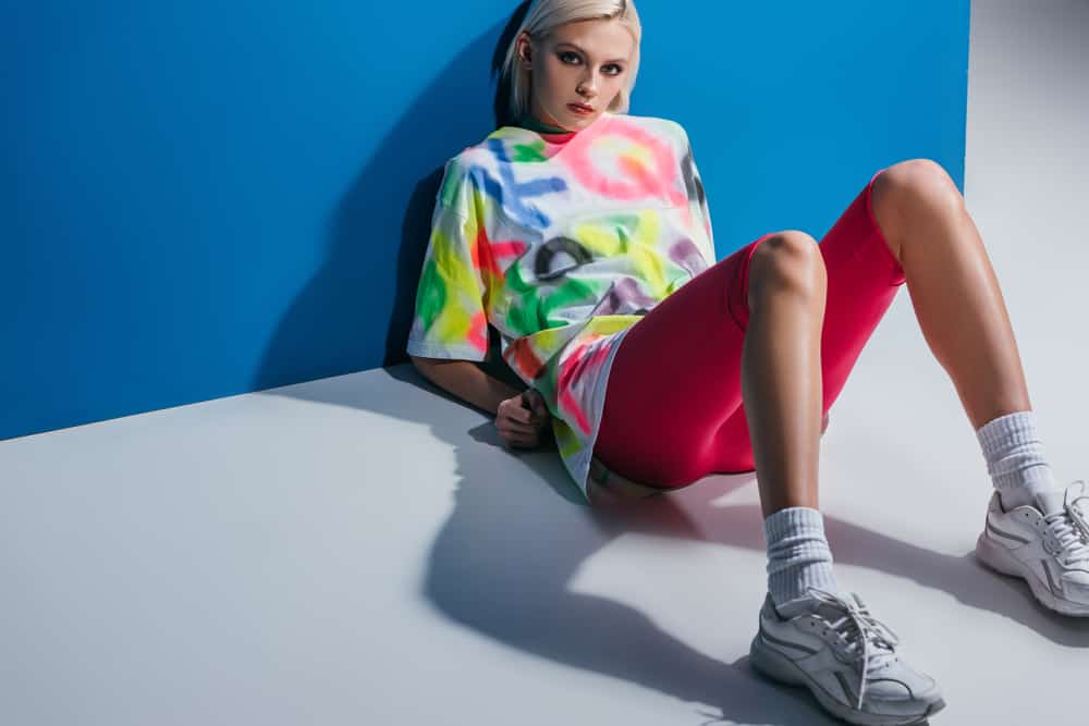 Model in a colorful graffiti shirt and pink bike shorts, poses against the blue wall.