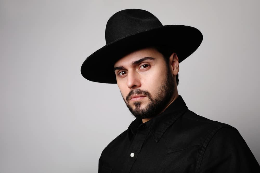 A bearded man wearing a black button shirt and a black fedora hat.