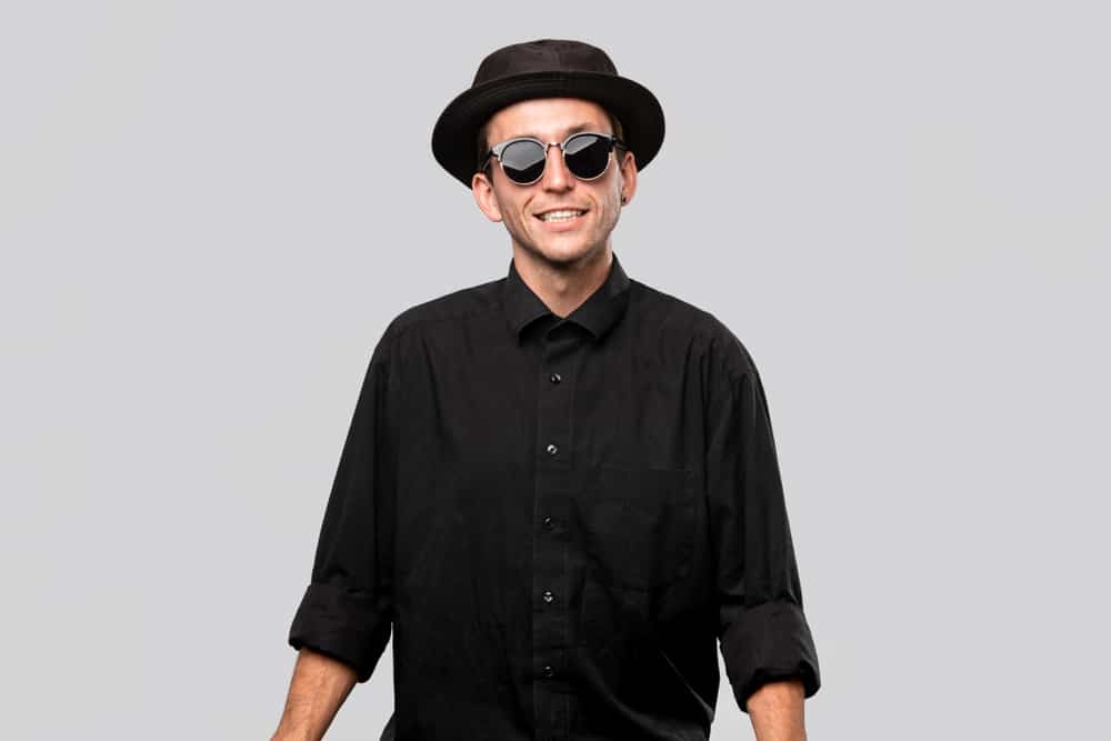 A man wearing a black button shirt with his black pork pie hat and sunglasses.