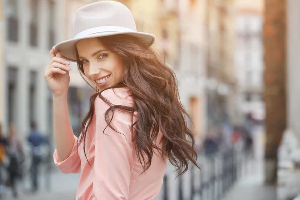 A woman with long wavy hair wearing a fedora and a pink blouse.