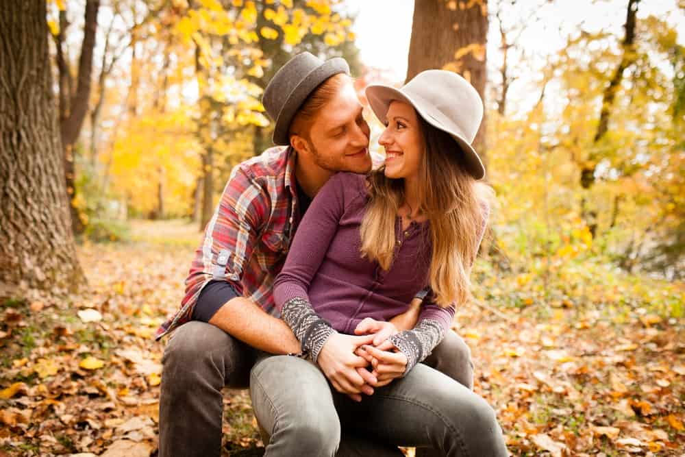 This is a couple in a forest during fall with the man wearing a pork pie hat.