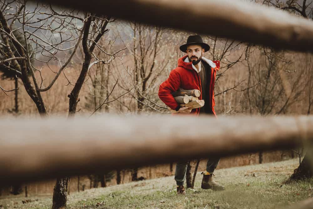 This is a bearded man in a ranch wearing a red jacket and a hat while collecting firewood.