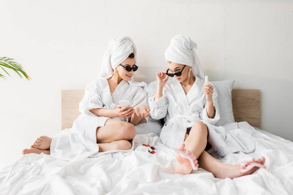 Women in bathrobes and sunglasses lying in bed.