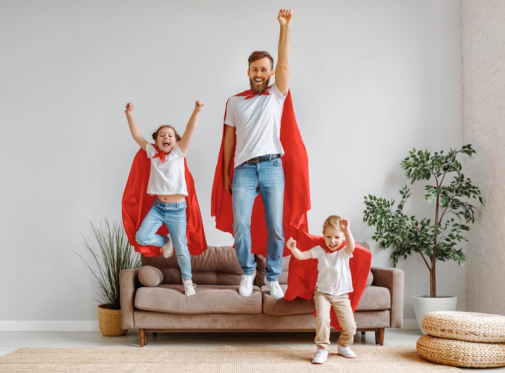Father with two children in casual clothes and red superhero cloaks jumping from couch to floor.