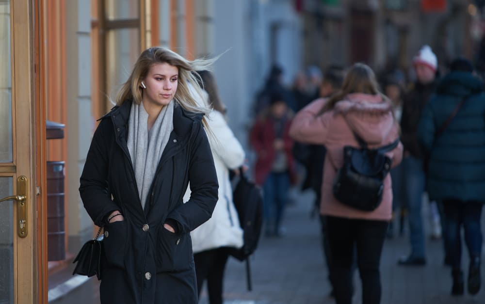 Blonde girl in black coat with headphones walking down the street.