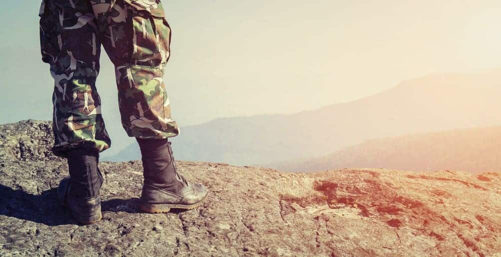 A soldier in uniform and combat shoes standing on the top of a mountain.