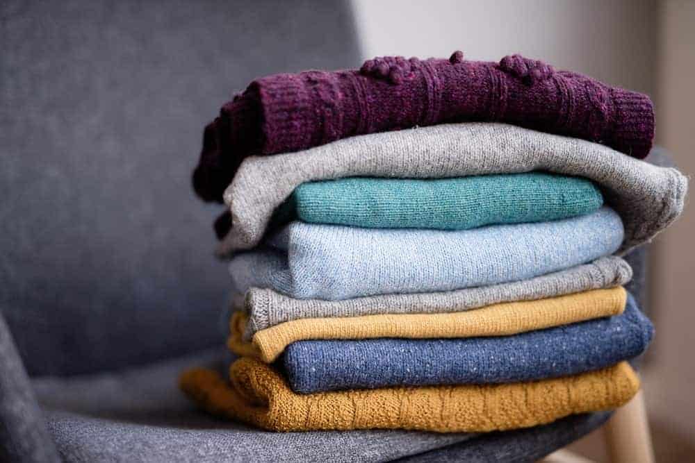 Knitted sweaters on pile above a gray chair.