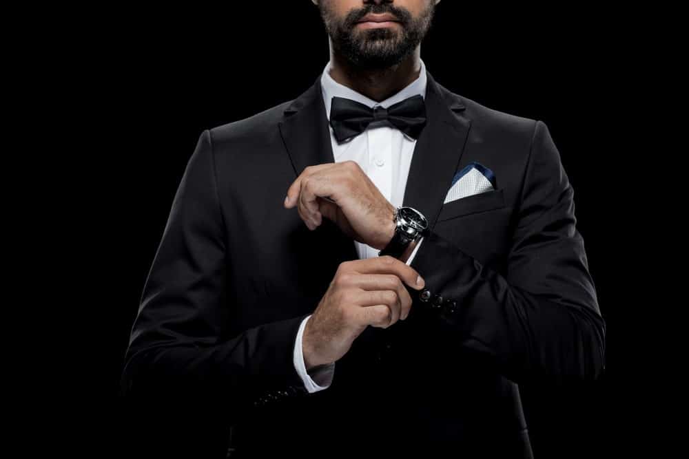 Cropped image of a man in a black tuxedo, tie, and wristwatch against a black background.