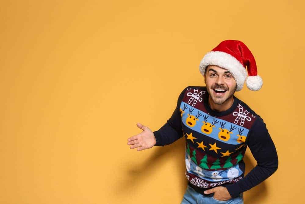 Man against an orange backdrop wearing a Christmas hat and an ugly Christmas sweater.