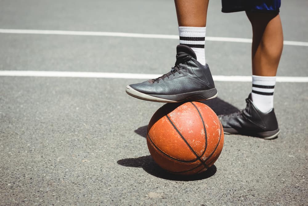 Man in mid-top basketball shoes standing with one leg on basketball.