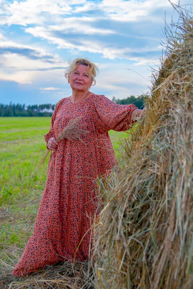 Elder woman with in a long country dress poses in haystacks.