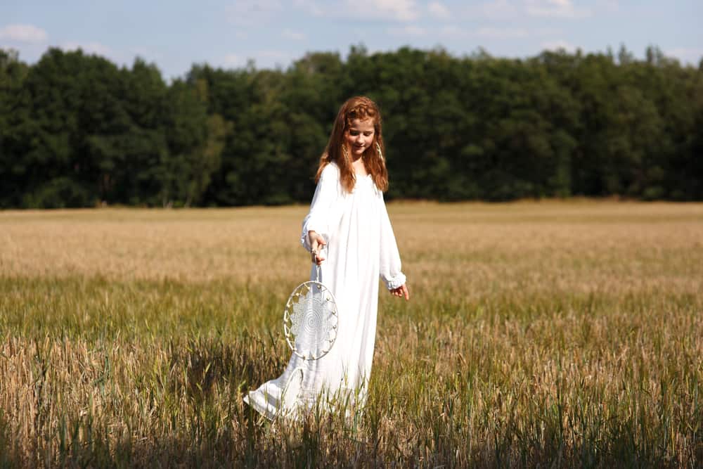 A girl in the middle of a field wearing a white peasant dress and holding a dream catcher.