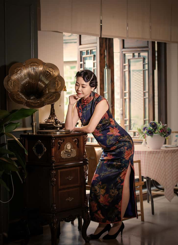 Woman in a qipao dress standing next to an ancient phonograph.
