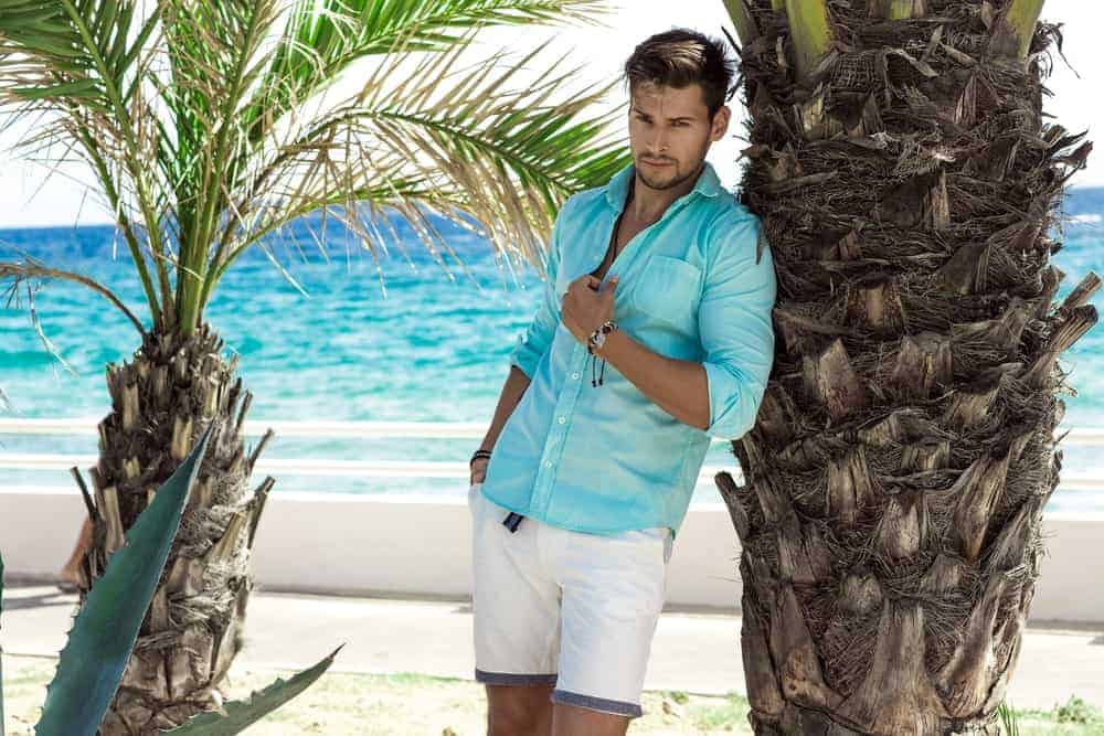 A man wearing a fashionable outfit of white shorts and blue button-down shirt at the beach.