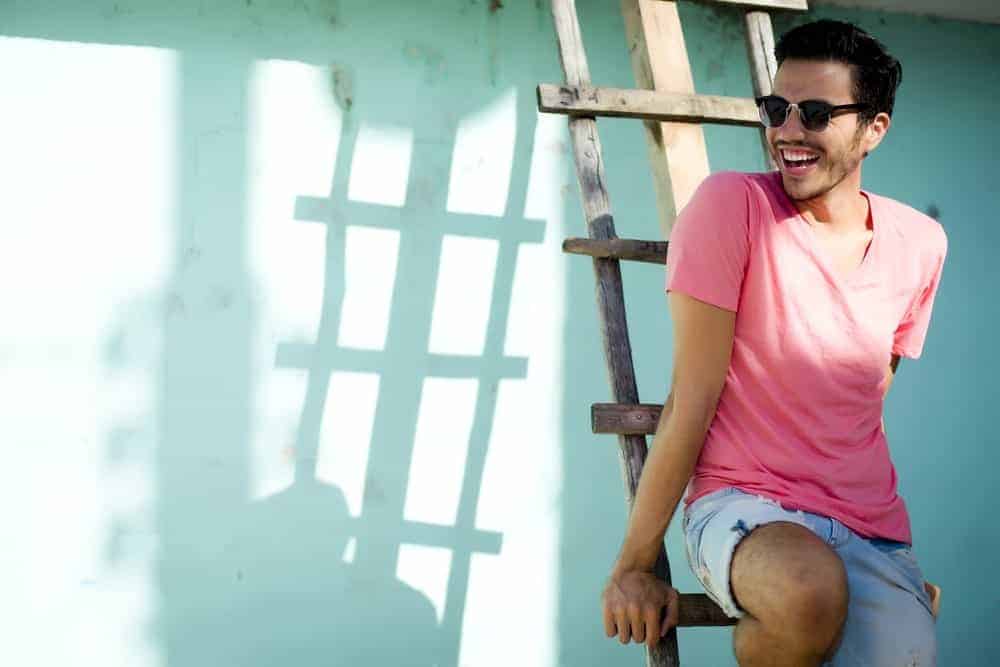 A man wearing a pair of sunglasses and a pastel pink shirt sitting on a wooden ladder.