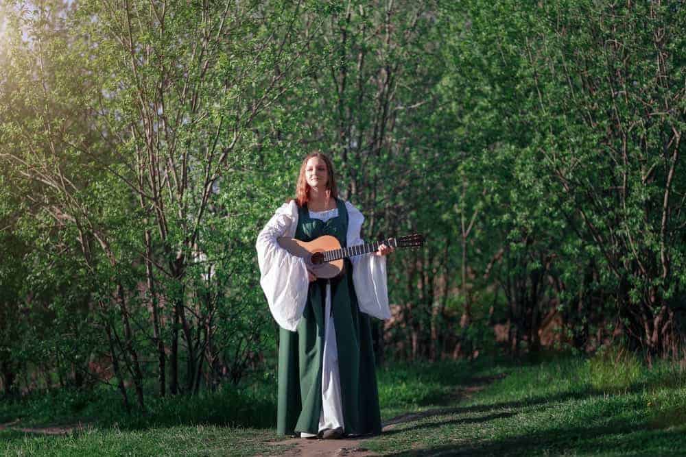 A woman playing the guitar while wearing a long medieval dress with peasant sleeves.