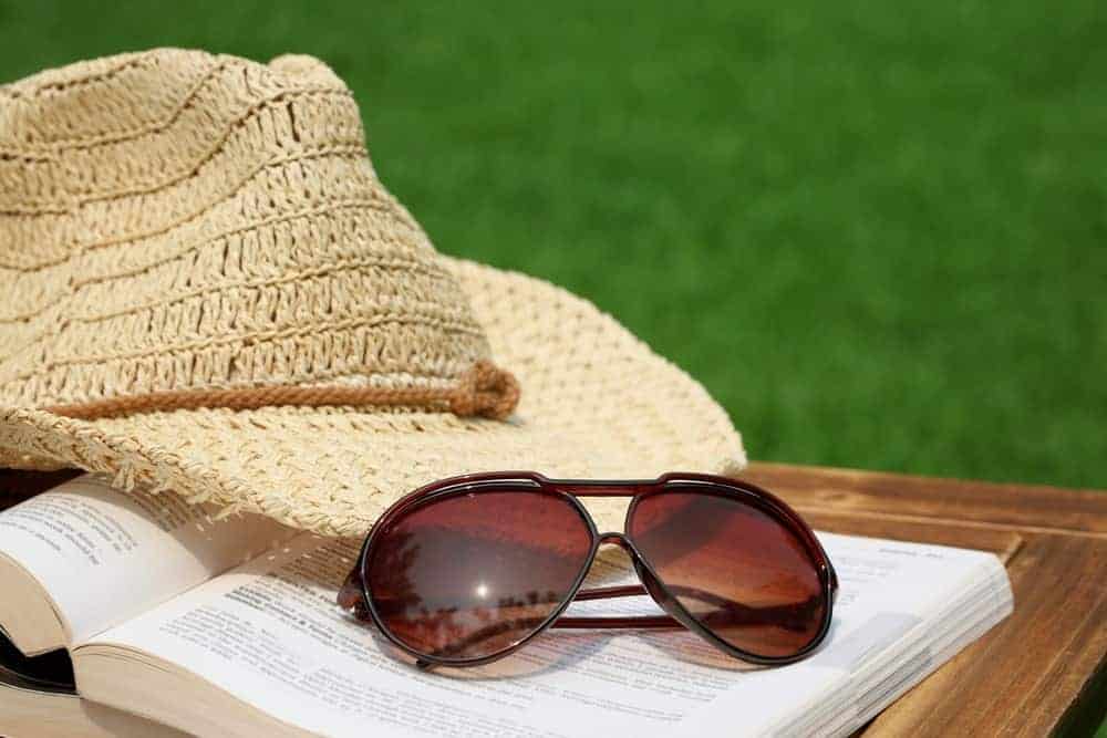 A pair of brown reading sunglasses with a book and a hat outdoors.