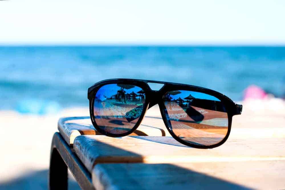 A pair of mirrored sunglasses on a wooden bench at the beach.