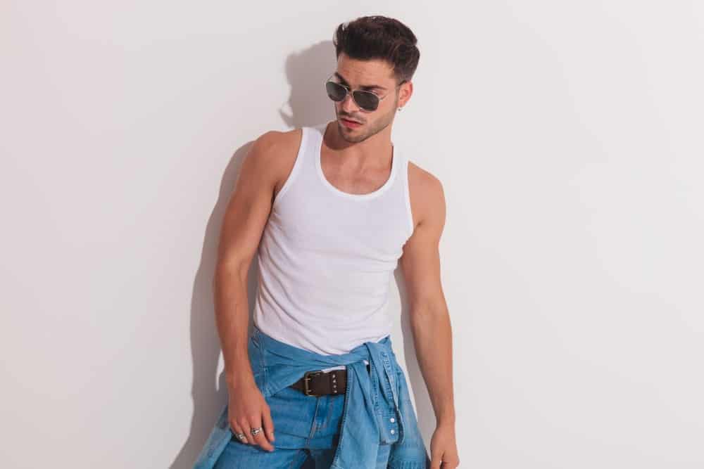 A man wearing a apir of shades, jeans and a white tank top undershirt.