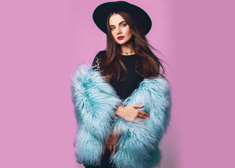 Woman in a stylish winter fluffy blue coat and black hat posing against a pink backdrop.