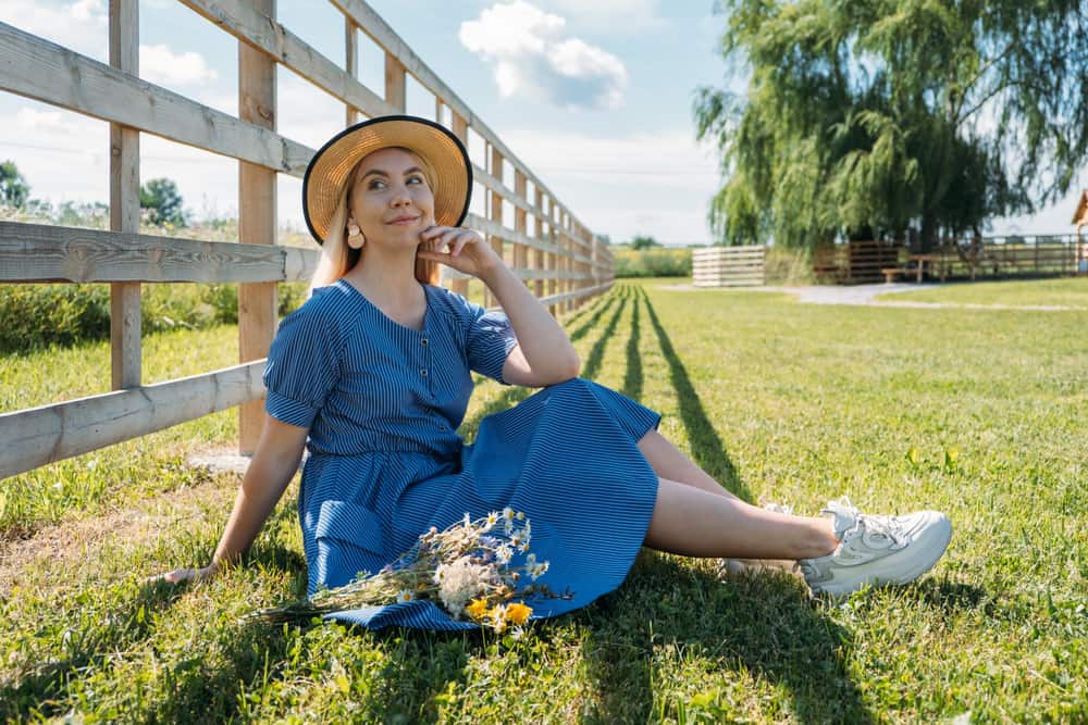 Woman in a straw hat, blue dress and sneakers sitting on a lawn near the fence.