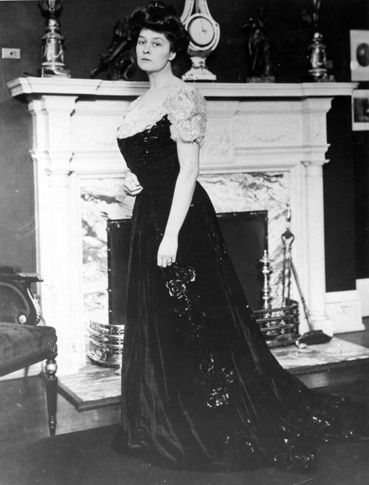 Woman in a gibson girl outfit standing near a fireplace.