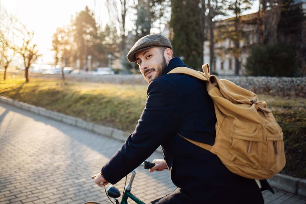 A man on a bike wearing a backpack and a brixton hat.