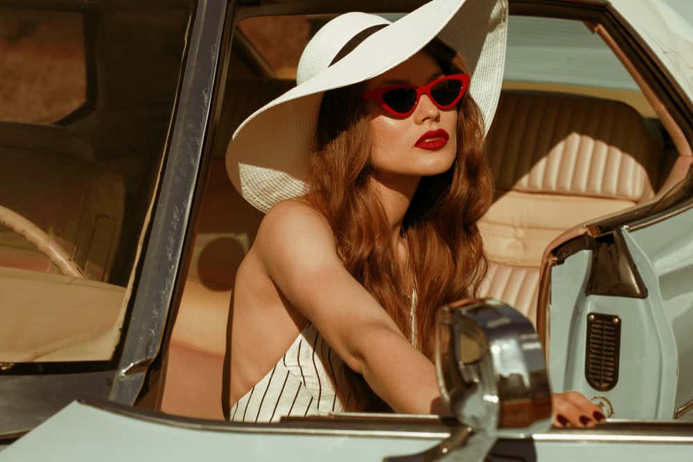 A woman wearing a wide brim hat and sunglasses in a vintage car.