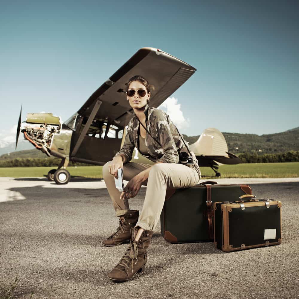Woman in military inspired outfit sitting on a suitcase against an airplane backdrop.