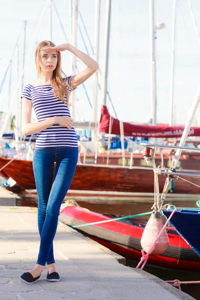 Girl in a striped shirt and denim pants standing near yachts.