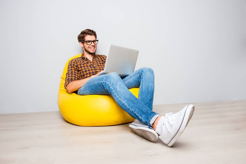 Man in checkered polo sitting on a yellow pouf while he uses his laptop.