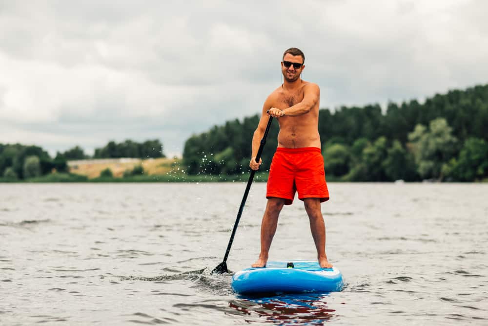 A man wearing a pair of red fitness board shorts while paddle boarding.