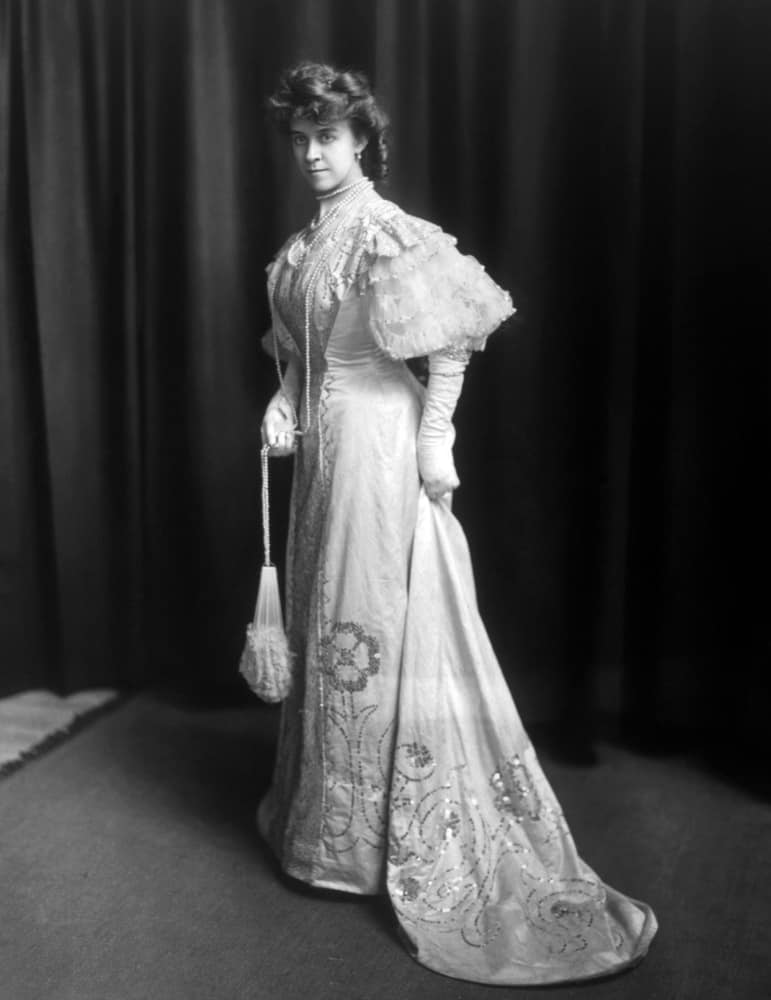 Woman wearing an 1890s wedding dress paired with layered necklace and a purse.