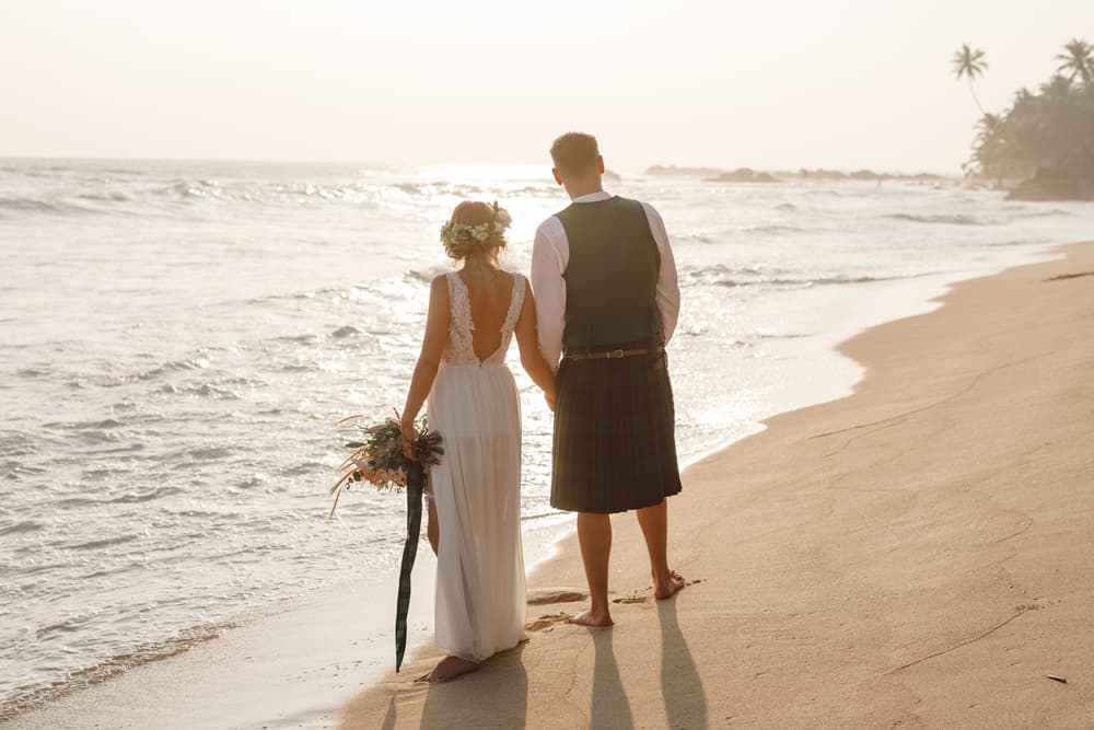 The groom wearing a kilt and the bride wearing a dress at the beach.