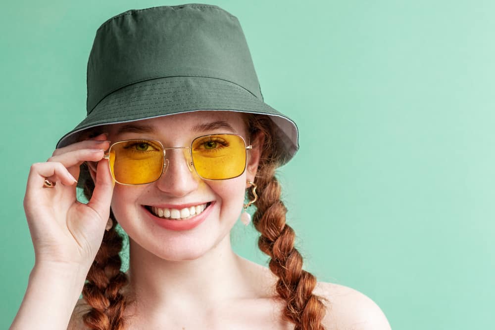 This is a woman with braids wearing a yellow pair of sunglasses and a green bucket hat.