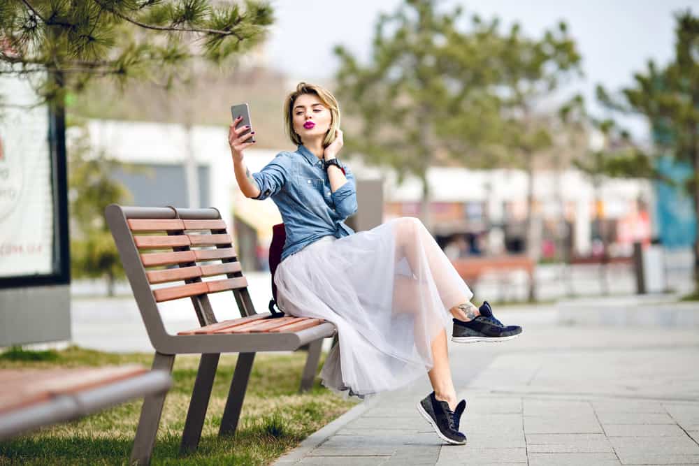 A woman taking a selfie on a park bench wearing a denim top and a white sheer skirt.