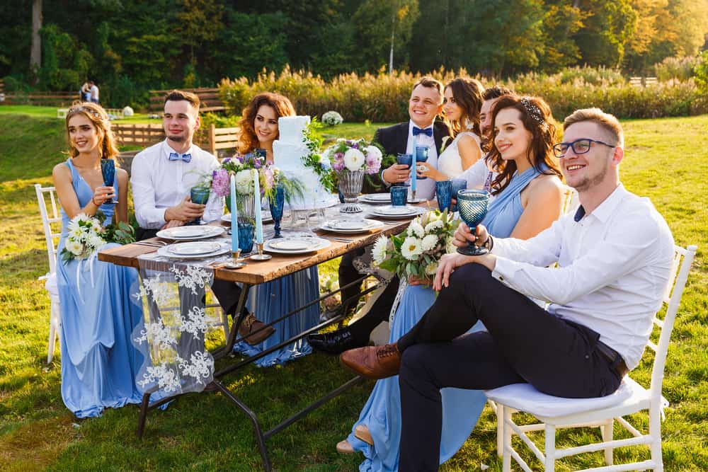 Newlyweds dining on the grass lawn with their groomsmen and bridesmaids who are wearing matching dresses.